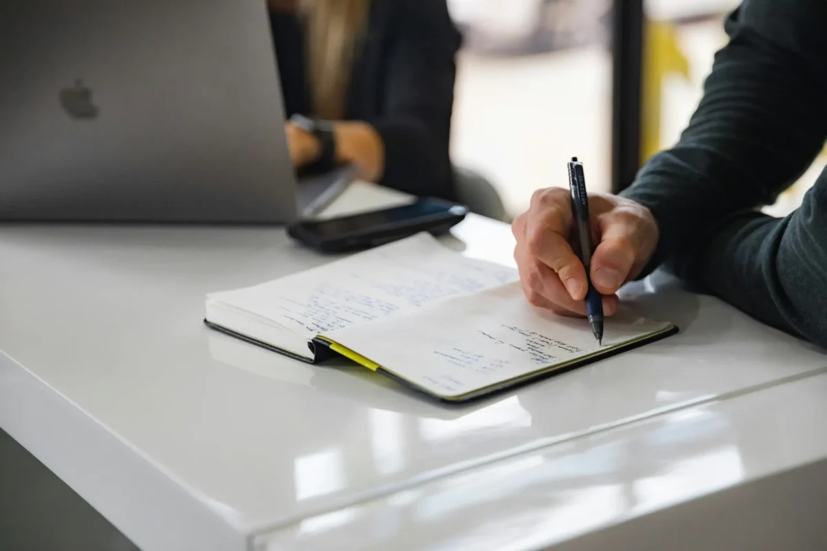 Person writing in a notebook beside a laptop during a financial planning meeting