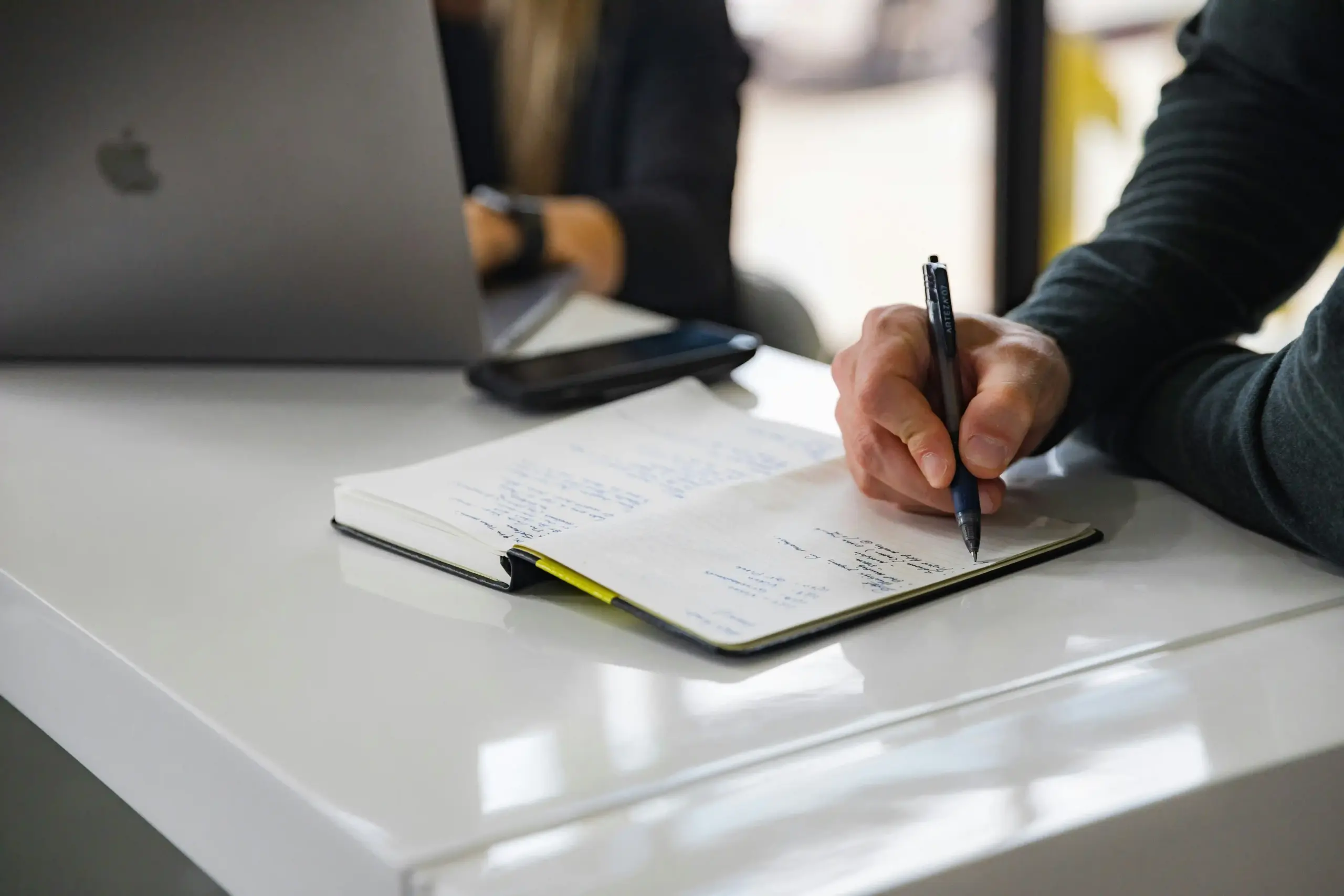 Person writing in a notebook beside a laptop during a financial planning meeting